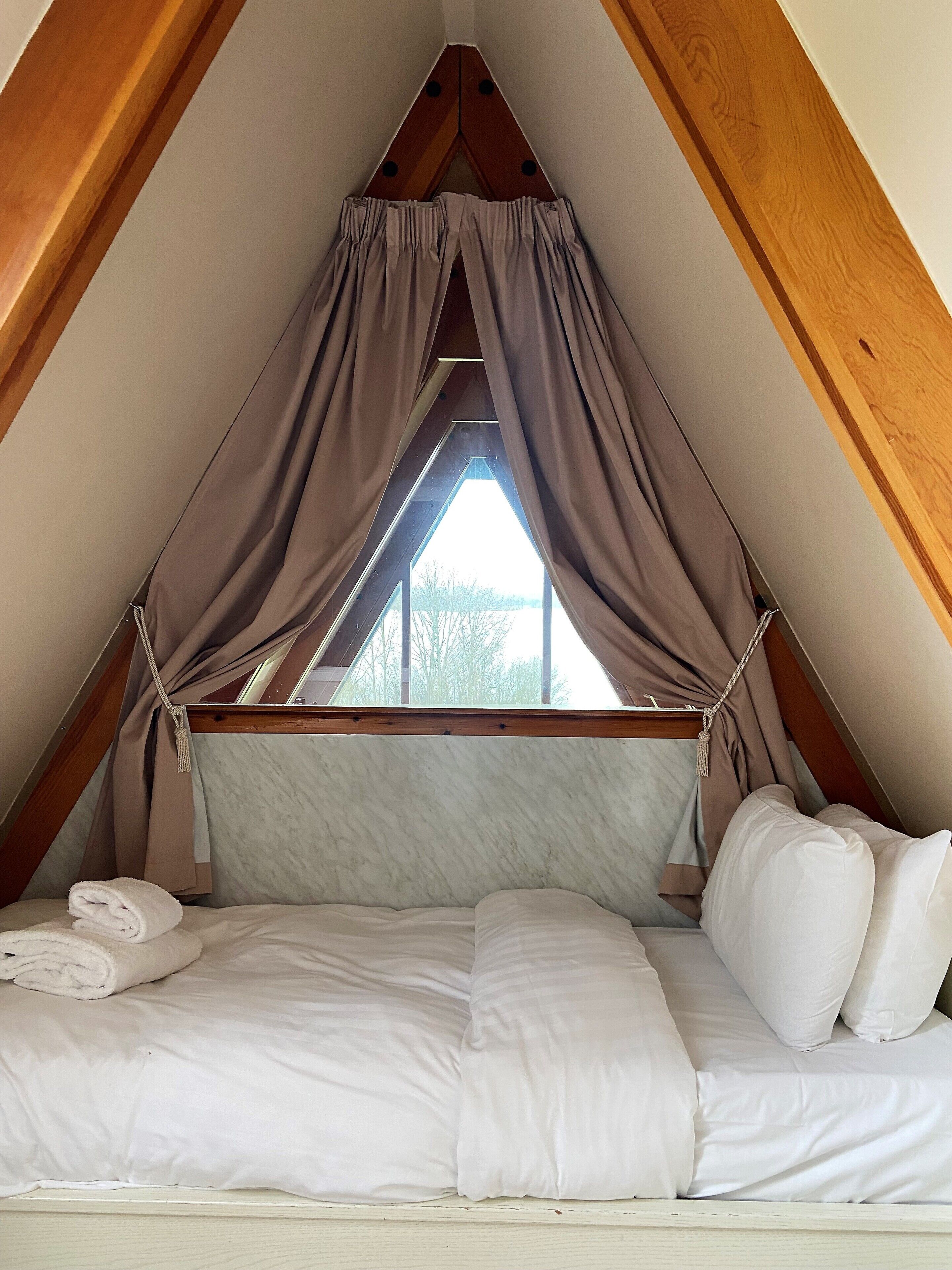 A cozy attic bedroom with a bed, a window, and a wooden ceiling.