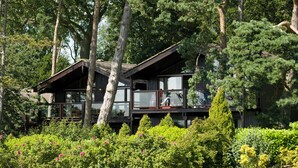 A two-story wooden cabin with large windows and a balcony, surrounded by dense greenery and trees. - Rutland Hall Lakeside Lodges (Oakham)
