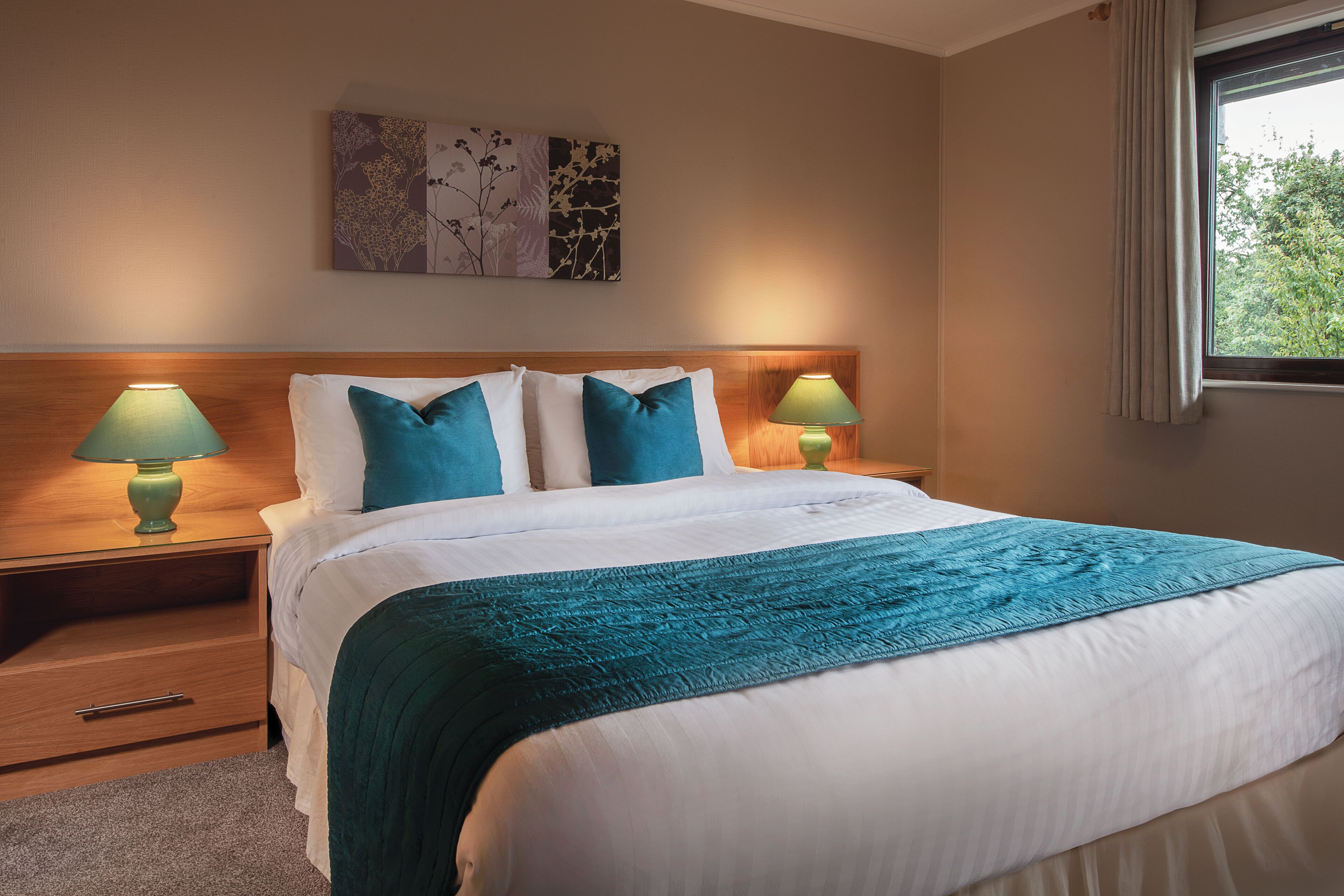 Un lit bien fait, agrémenté d’un plaid turquoise, de deux coussins assortis et de deux lampes de chevet.