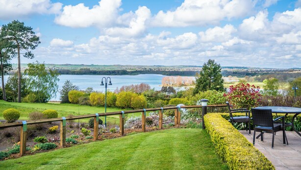 A patio with a view of a lake, surrounded by well-maintained hedges and a variety of plants. - Rutland Hall Lakeside Lodges (Oakham)