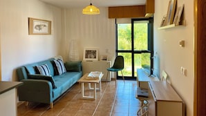 A modern living room with a sofa, a coffee table, and a desk. There is a window with a view of greenery outside. - Apto Las Dunas de Langosteira en Finisterre (finisterre)