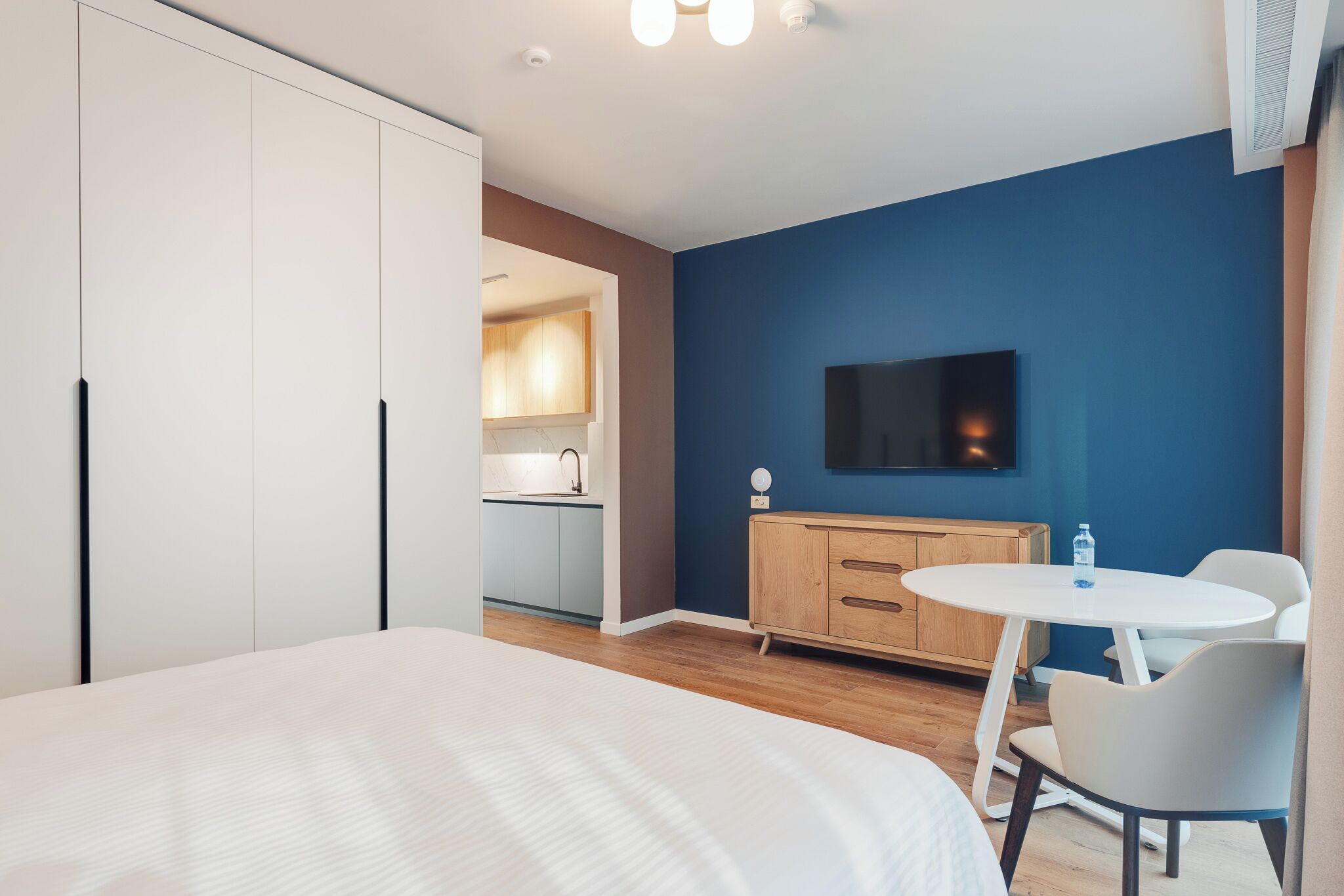 A modern hotel room with a bed, a small dining table, a TV, and a kitchenette in the background.