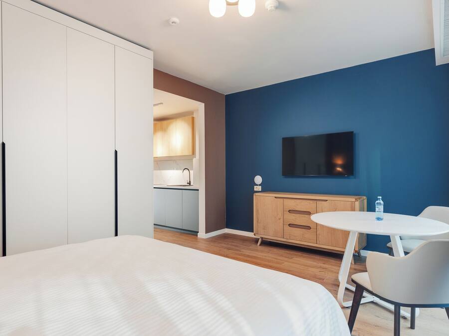 A modern hotel room with a bed, a small dining table, a TV, and a kitchenette in the background.