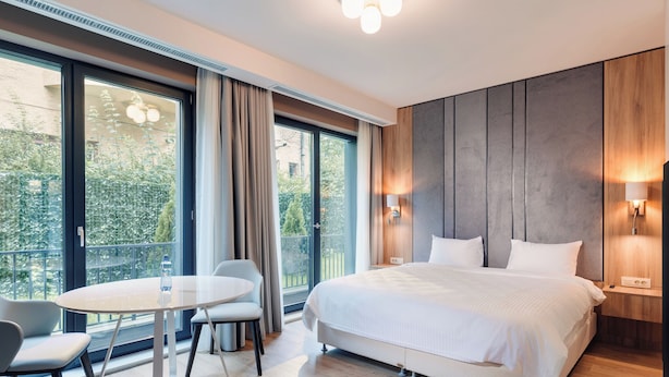 Une chambre d’hôtel moderne avec un grand lit, une table à manger ronde et un balcon offrant une vue sur la verdure.
