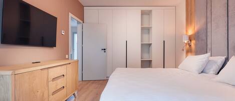 A modern bedroom with a large bed, a TV mounted on the wall, a wooden dresser, and built-in shelving.