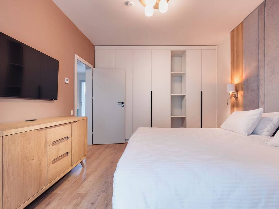 A modern bedroom with a large bed, a TV mounted on the wall, a wooden dresser, and built-in shelving.