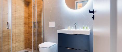 A modern bathroom with a glass shower enclosure, a white toilet, a dark vanity with a mirror, and a sink with toiletries.
