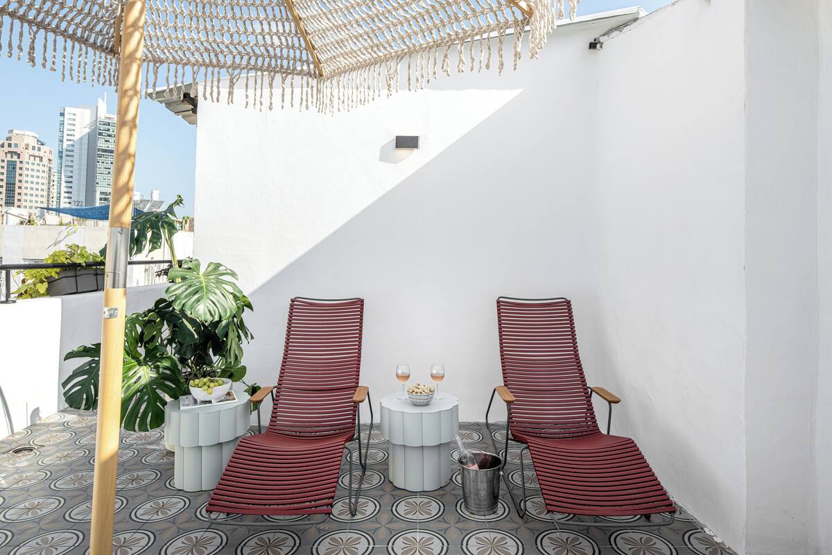 A rooftop terrace with two red-striped chairs, a small round table with a bowl of fruit and two glasses, and a potted plant.