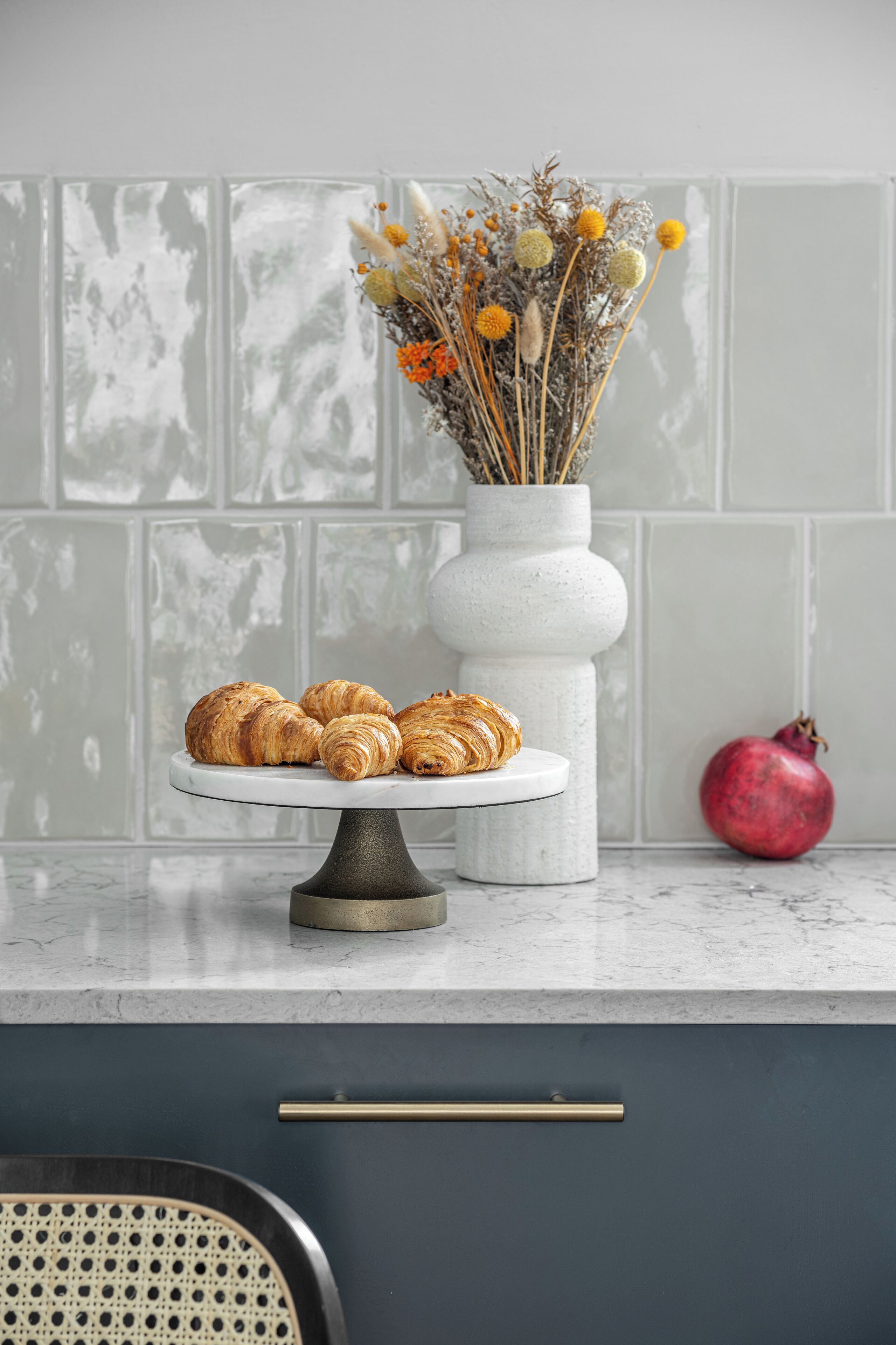 A kitchen counter with a white vase containing dried flowers and a pomegranate, a white cake stand with croissants, and a wicker chair.