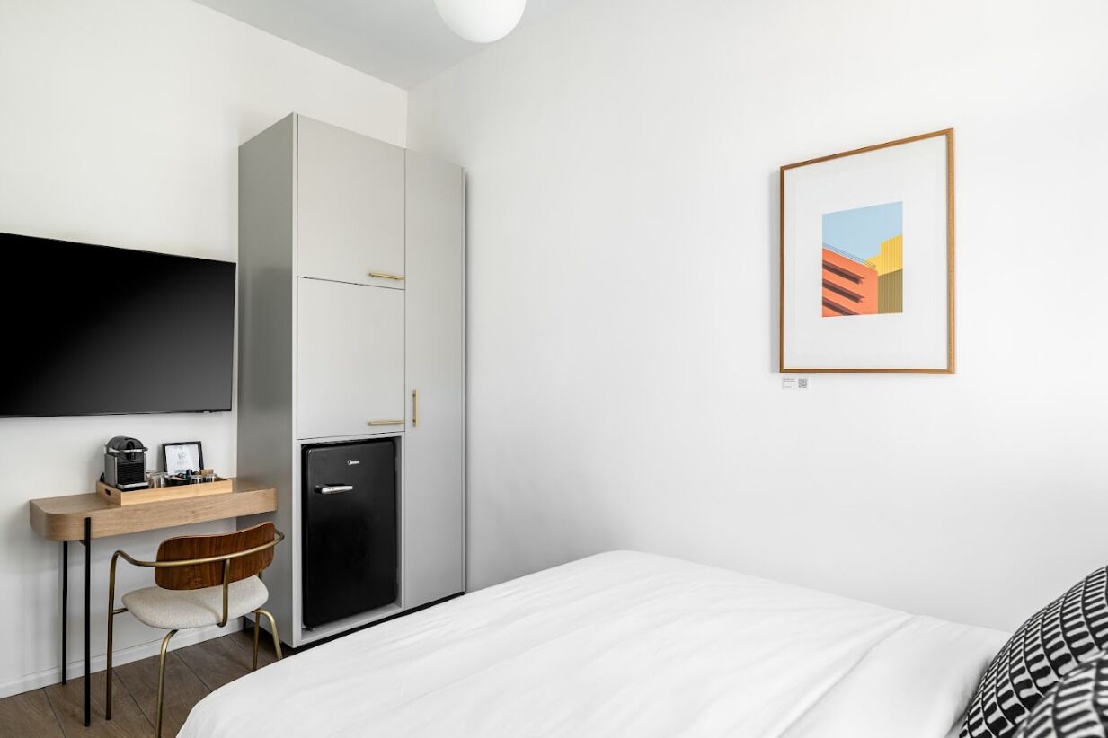 A modern hotel room with a bed, a desk with a chair, a refrigerator, a flat-screen TV mounted on the wall, and a framed picture on the wall.