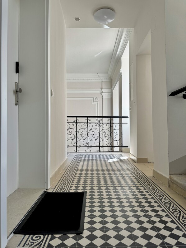A hallway with a checkered floor, a black mat at the entrance, and a white door on the left.