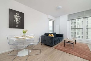 A modern living room with a dark grey sofa, a glass dining table with chairs, a coffee table, and a large window with a city view.