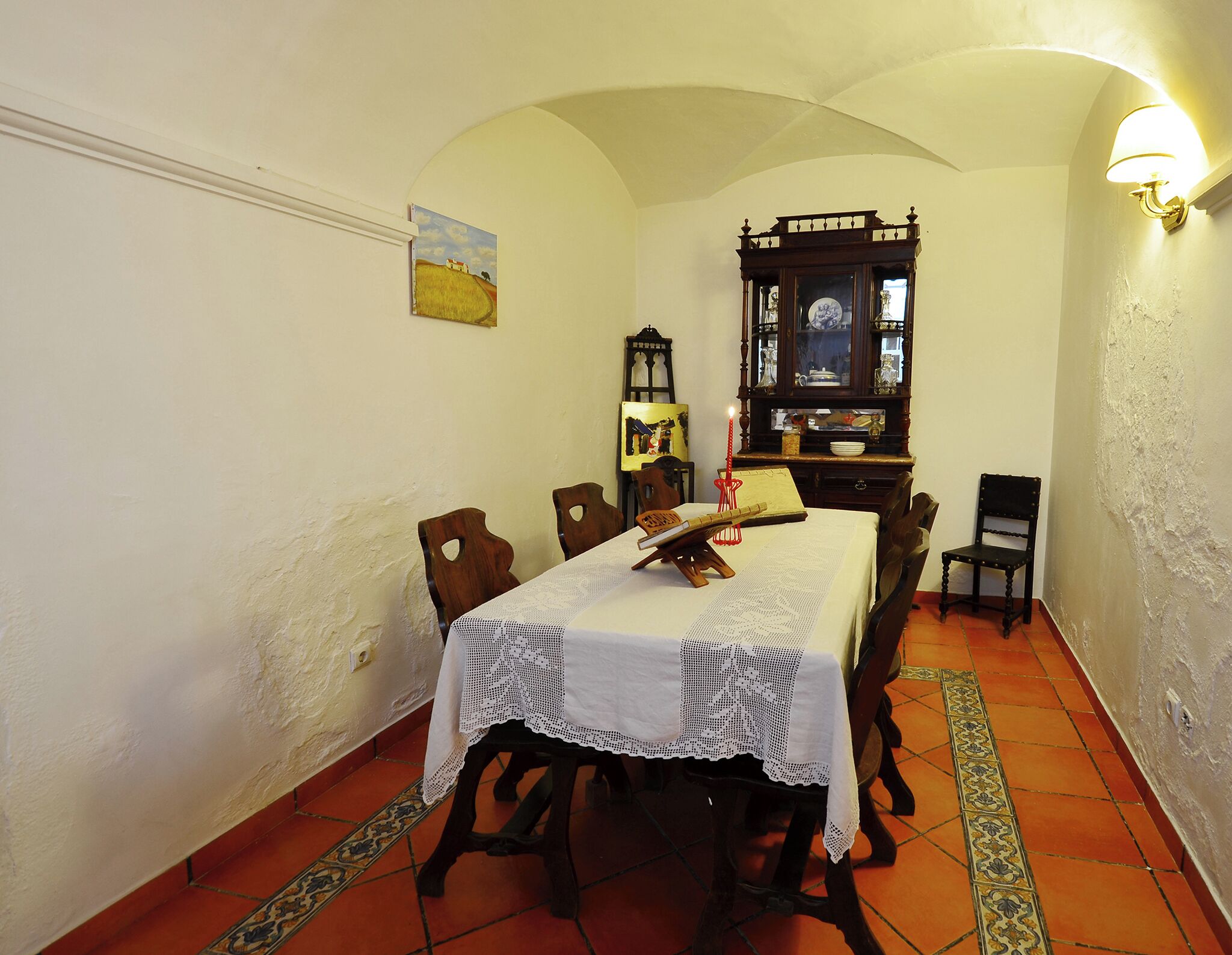 A dining room with a table set for four, surrounded by wooden chairs. A wooden cabinet with glass doors is against the wall, and a painting hangs above the table.