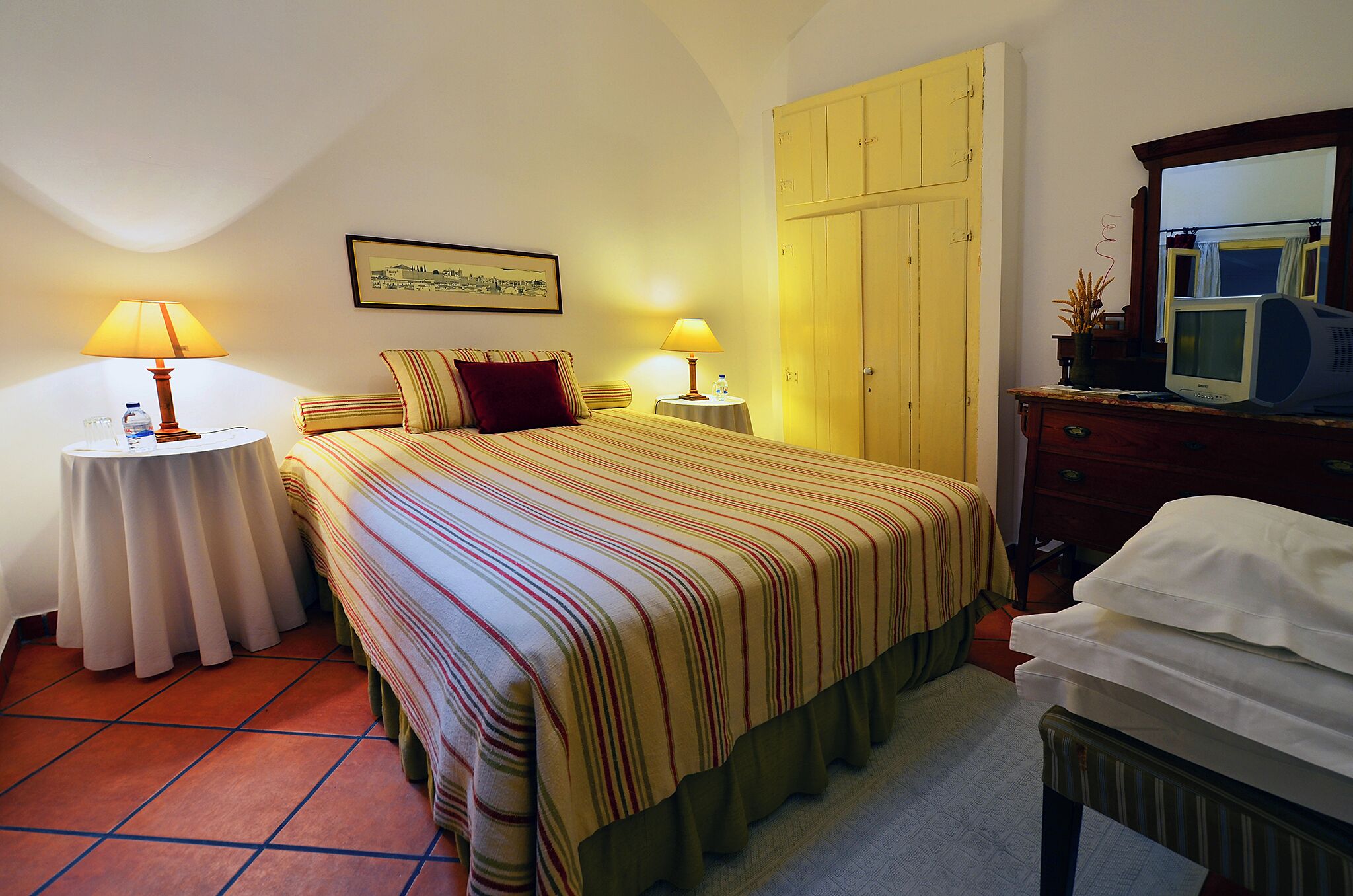 Ein Hotelzimmer mit einem Bett, zwei Nachttischen mit Lampen, einem kleinen Tisch mit Pflanze, einem Kleiderschrank und einem Spiegel mit Fernseher.