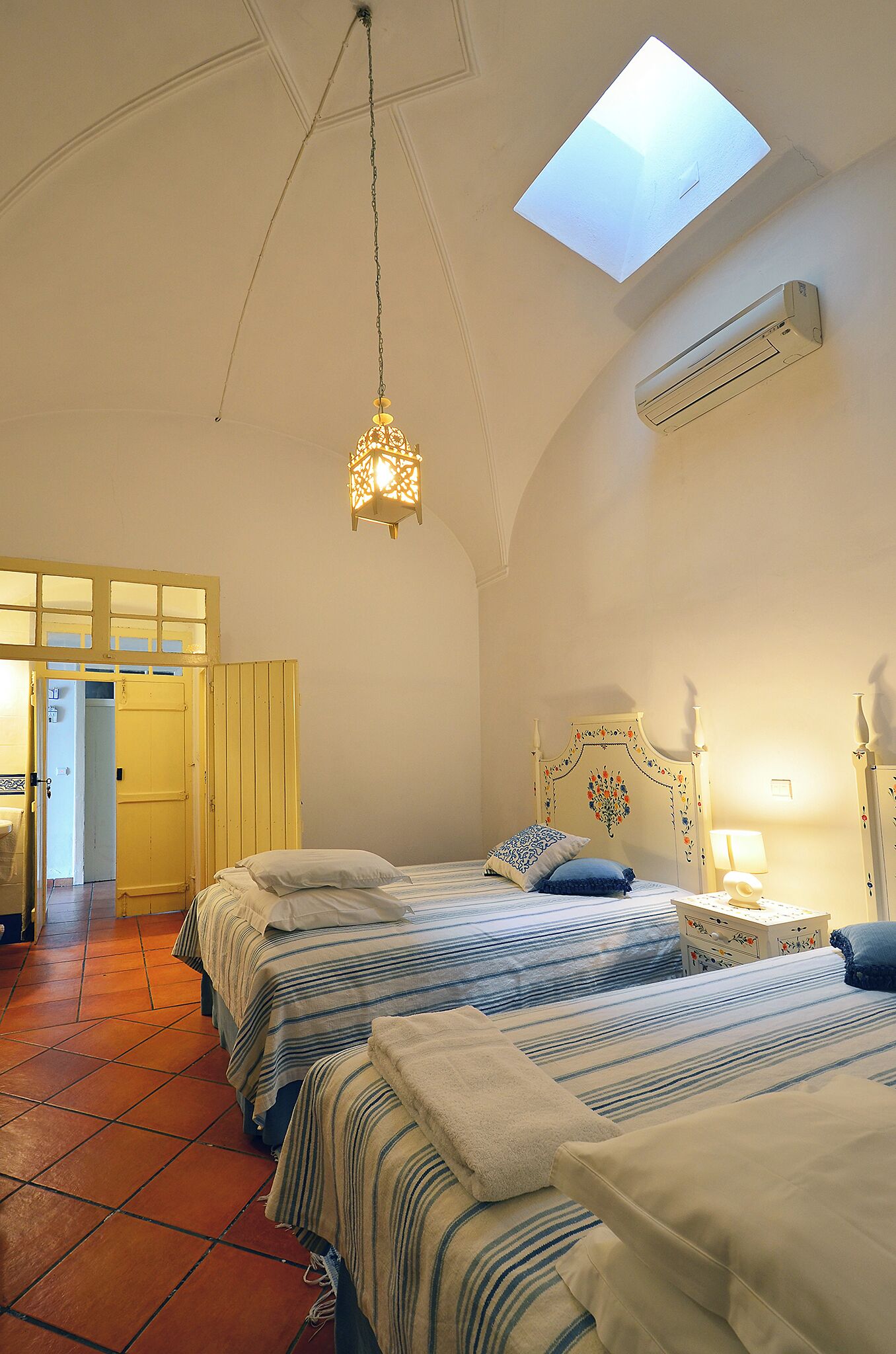 A bedroom with two beds, a skylight, an air conditioning unit, and a bedside table with a lamp.