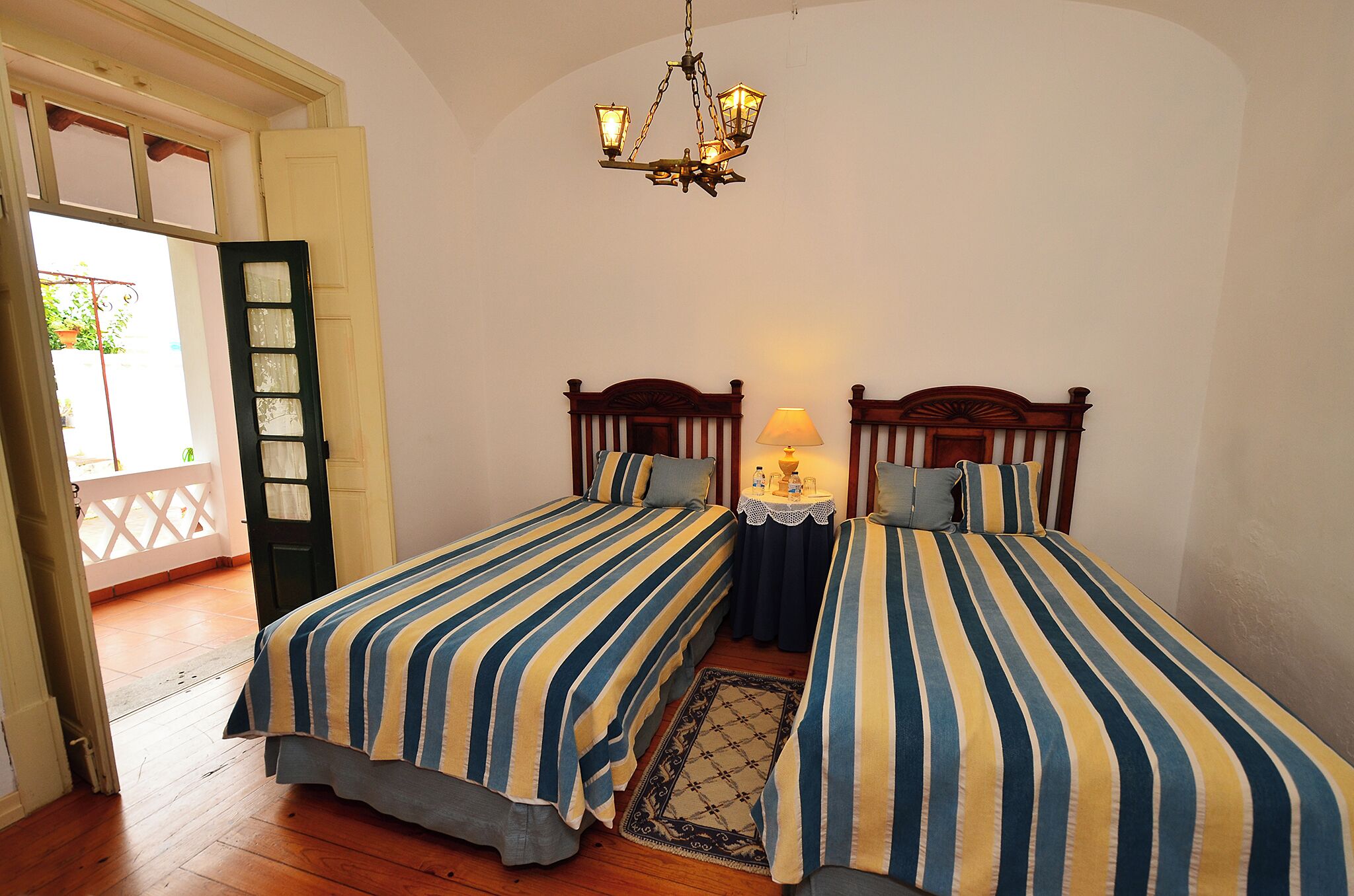 A room with two beds, a chandelier, a small table with a lamp, and a patterned rug.