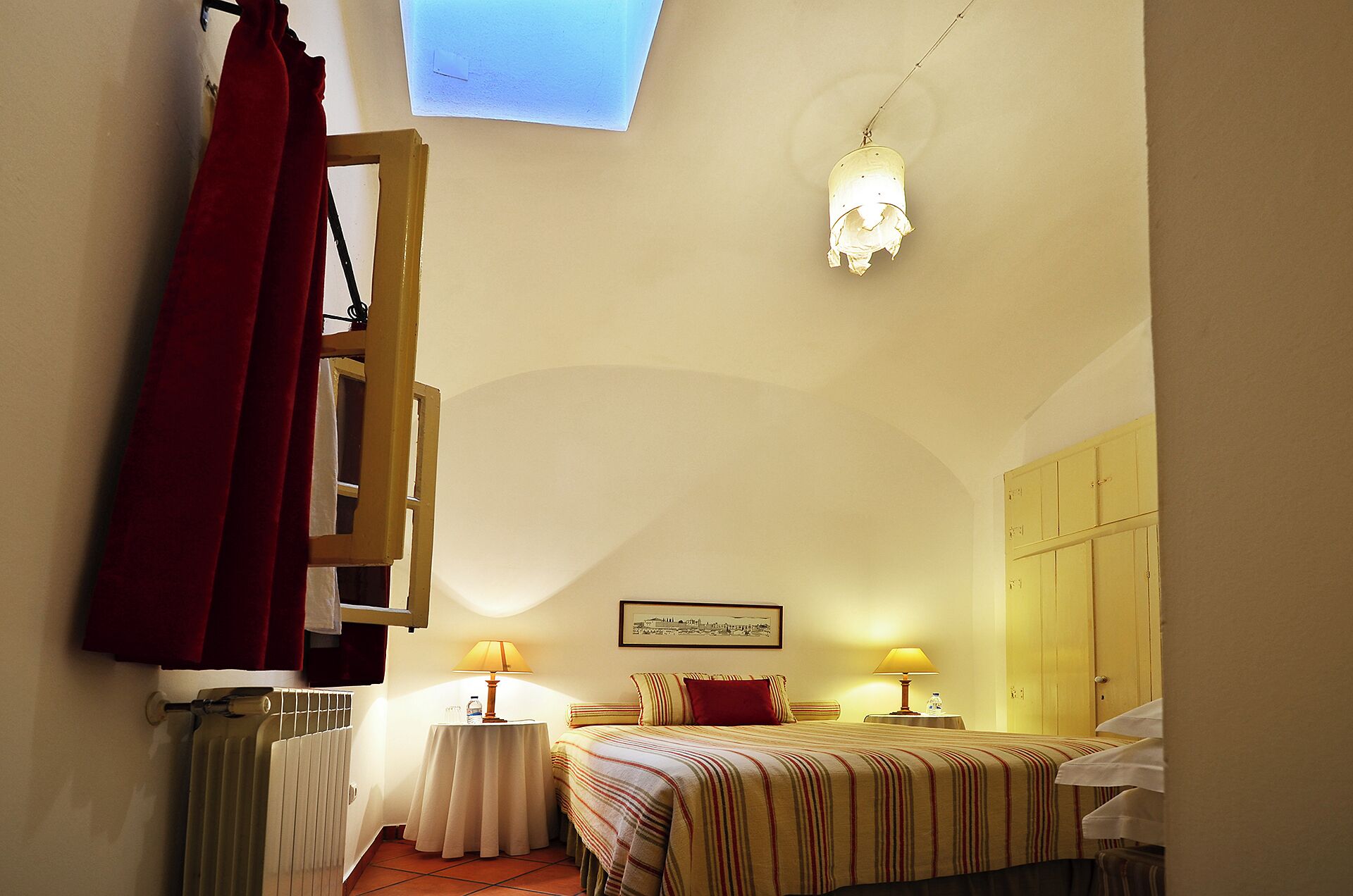 Ein Hotelzimmer mit einem Bett, zwei Nachttischen mit Lampen, einem Heizkörper, einem Fenster mit Vorhängen, einer Pendelleuchte und einem gerahmten Bild an der Wand.