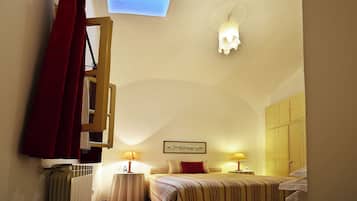 Une chambre d’hôtel comprenant un lit, deux tables de chevet avec des lampes, un radiateur, une fenêtre avec des rideaux, un luminaire suspendu et un tableau encadré au mur.