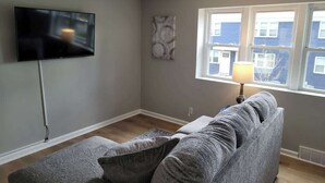 A living room with a grey sofa, a flat-screen TV, a floor lamp, and a window with a view of buildings.