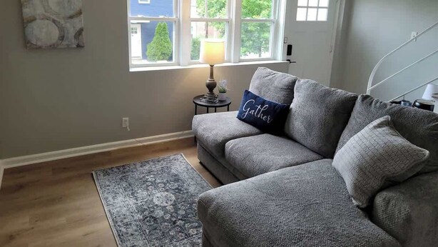 A living room with a grey sofa, a rug, a lamp, and a window with a view of a house and trees.