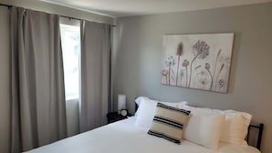 Una cama bien hecha con sábanas blancas y almohadas a rayas, una mesita de noche con lámpara y reloj, y un cuadro colgado en la pared con flores.