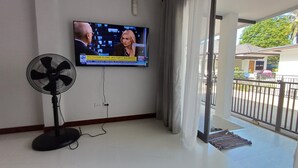 A room with a television mounted on the wall, a standing fan, and a view of the outside balcony.