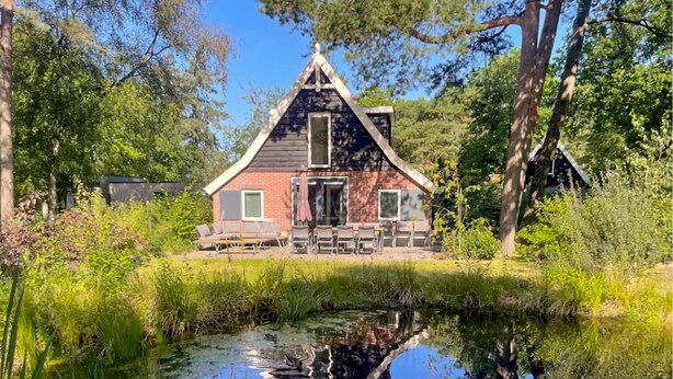 A rustic cabin with a sloped roof, a front porch with chairs, and a pond in the foreground.