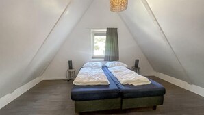A small, triangular attic bedroom with a bed, two bedside tables, a window with curtains, and a hanging light fixture.
