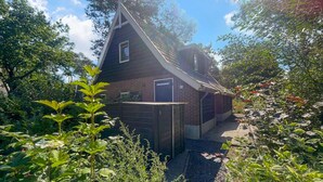 A small, dark wooden house with a sloped roof, surrounded by dense greenery and trees.