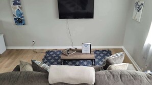 Ein Wohnzimmer mit grauem Sofa, einem Couchtisch mit Buch und Dekoration, einem an der Wand befestigten Flachbildfernseher und einem gemusterten Teppich.