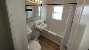 Ein Badezimmer mit Toilette, Waschbecken und Badewanne. Ein Fenster sorgt für natürliches Licht, über dem Waschbecken befindet sich ein Spiegel.