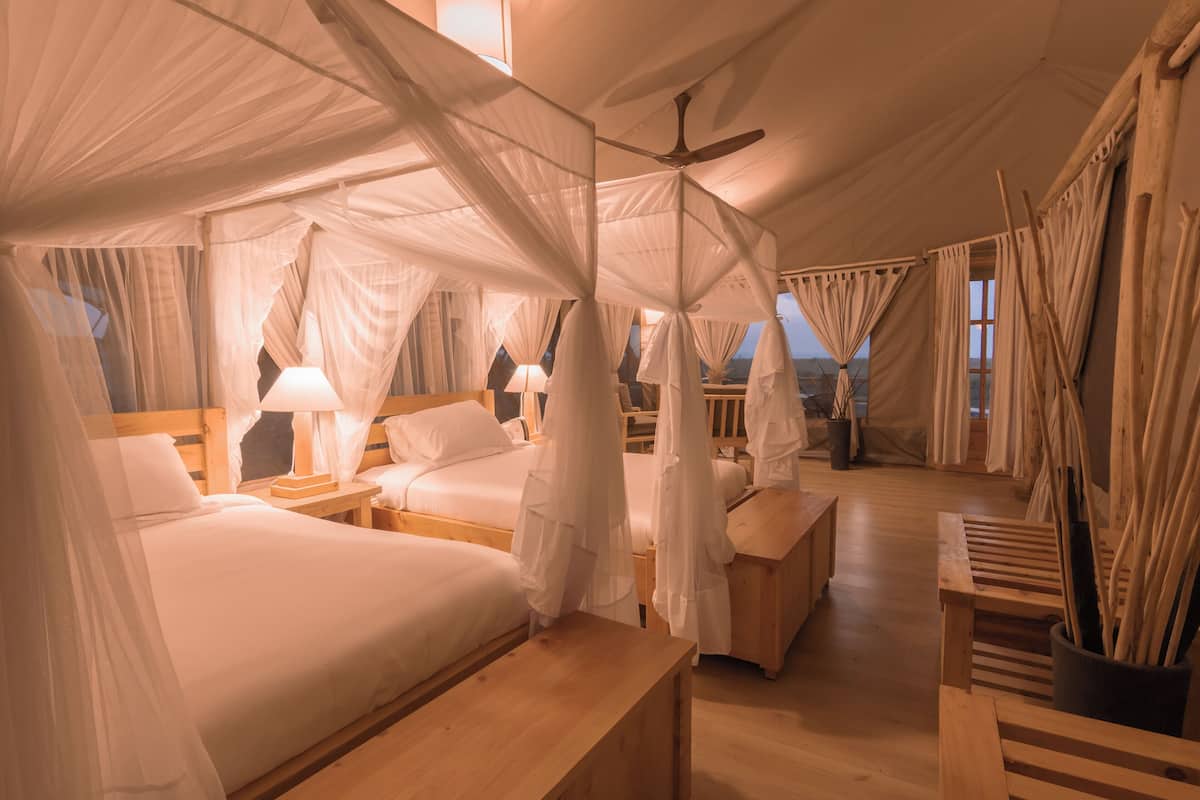 A tent-style room with four beds, each with white linens and canopy-style curtains. There are wooden nightstands with lamps, and a ceiling fan is visible.