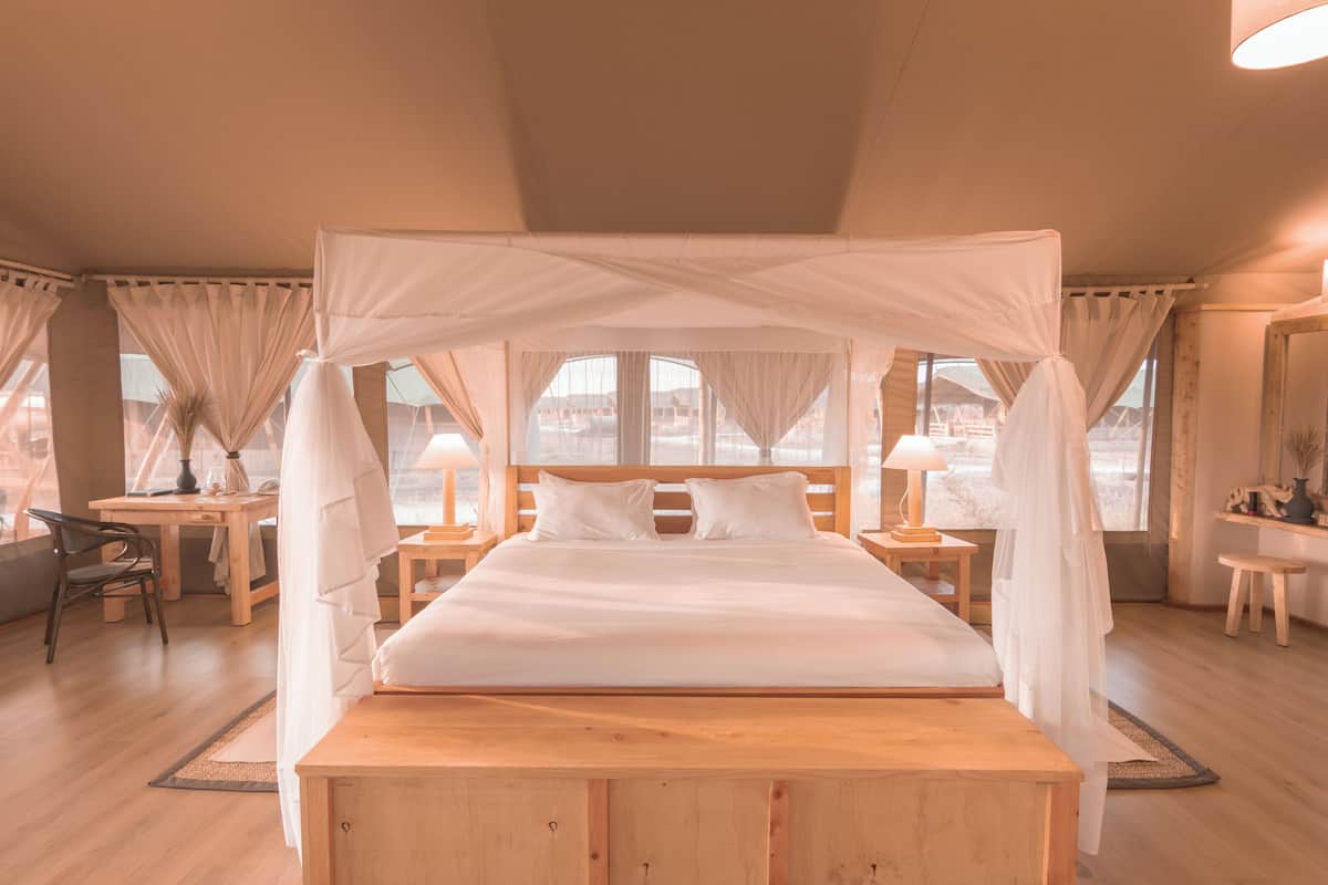 A bed with a canopy, wooden headboard, and white bedding. There are two bedside tables with lamps, a chair, and a desk with a lamp.