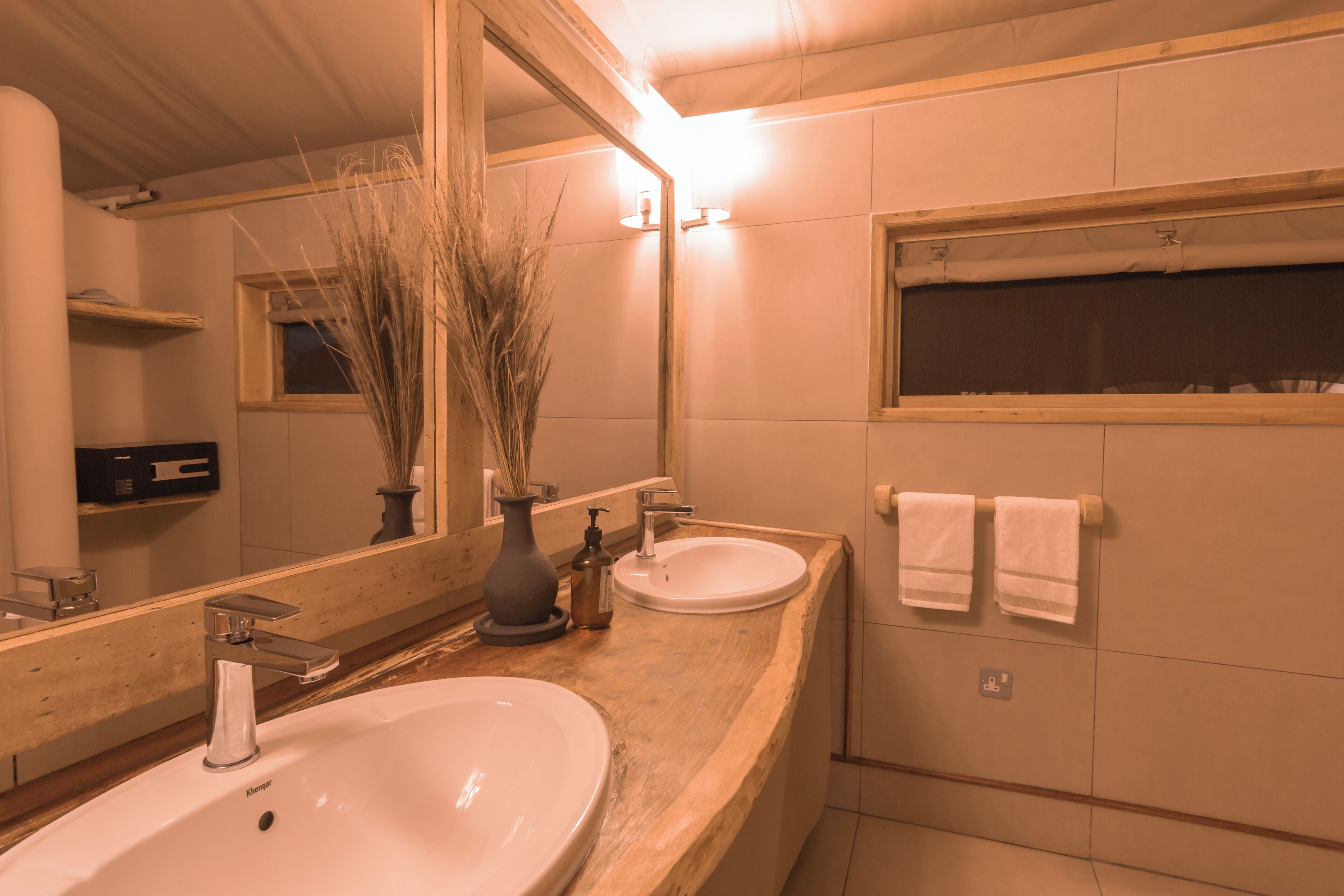 A bathroom with a wooden vanity, two sinks, a large mirror, and a window above the bathtub.