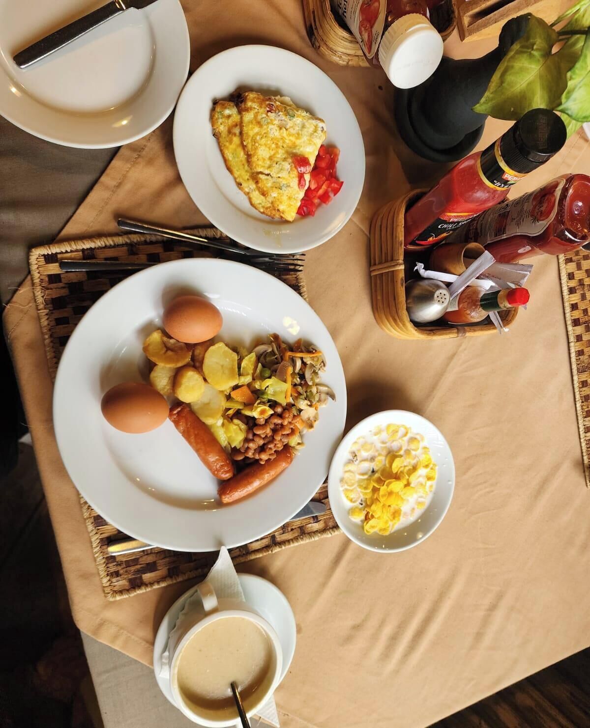 A breakfast setting with plates of eggs, sausages, and potatoes, a bowl of cereal, and a cup of coffee.
