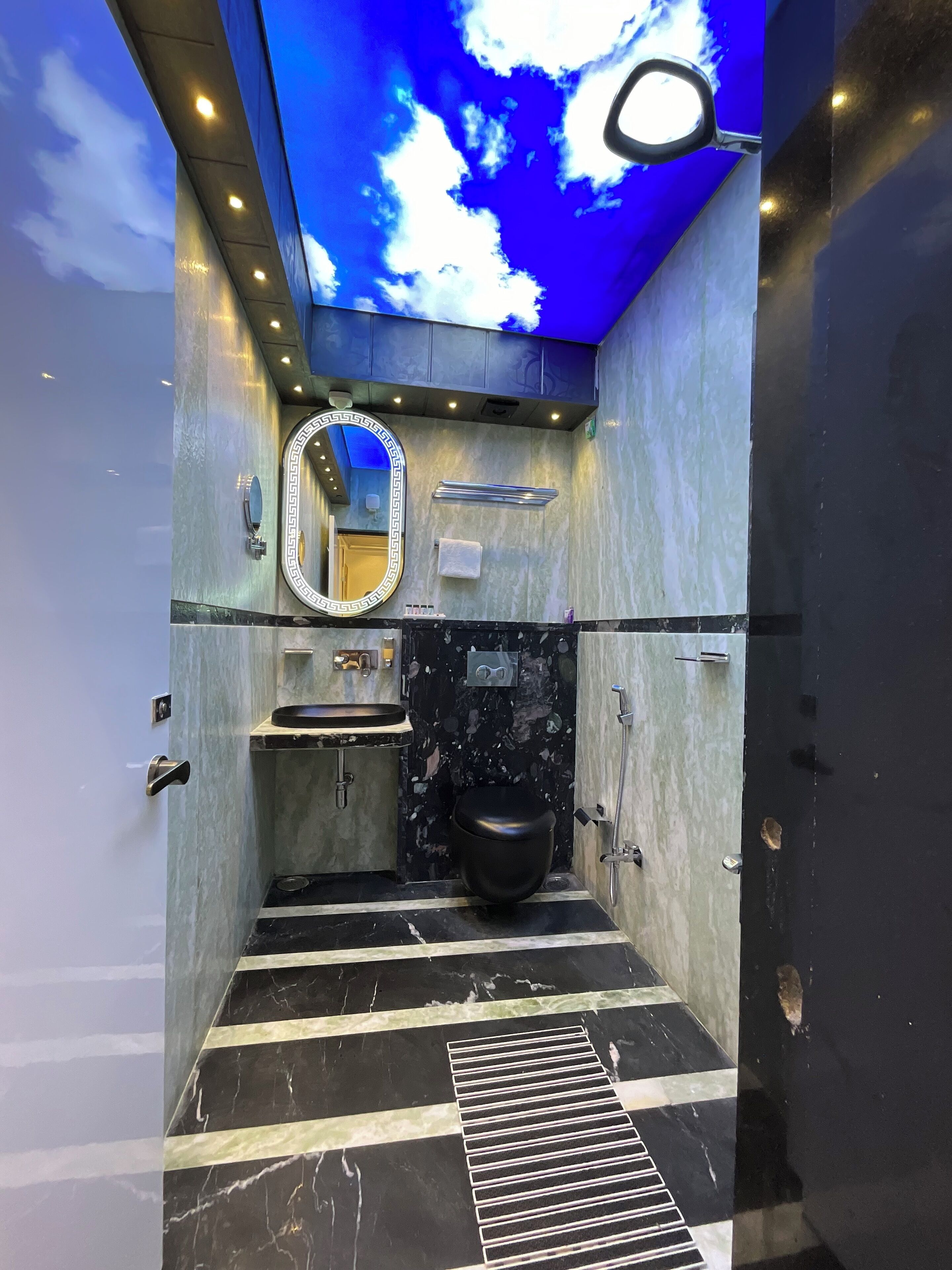Et moderne badeværelse med klinkegulv i marmor, et sort toilet, en vask med marmorplade, et spejl med dekorativ ramme og et loft med skyformet lysinstallation.