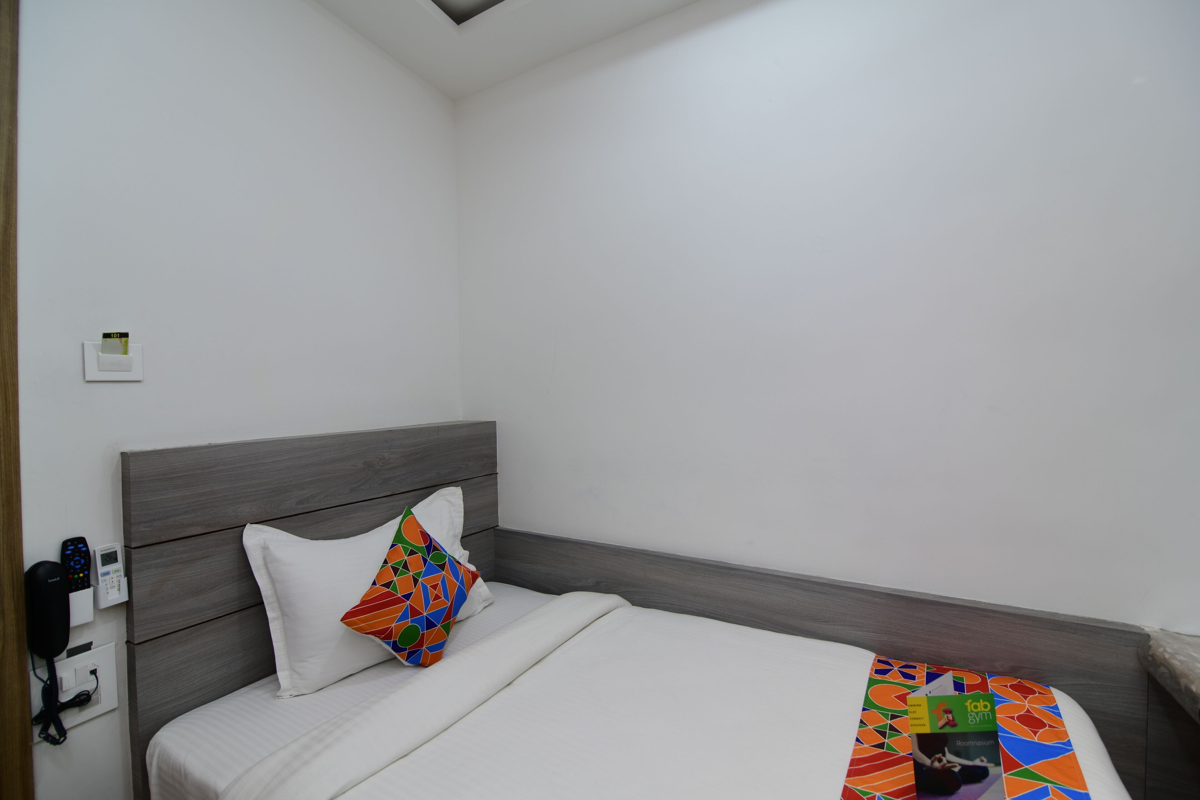 A neatly made bed with a white pillow and a colorful geometric pillow, a bedside table with a book, and a wall-mounted phone.