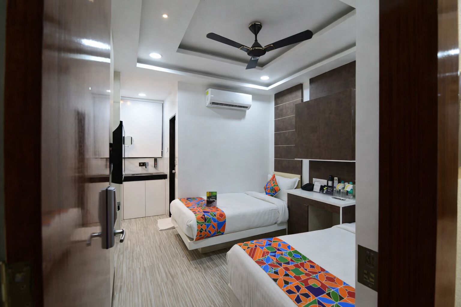 A hotel room with two beds, a ceiling fan, an air conditioning unit, a small kitchenette, and a bedside table with toiletries.