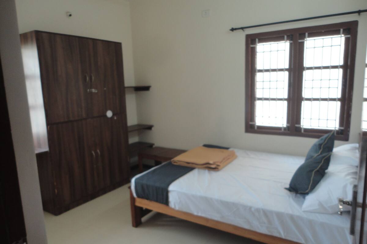 Une chambre avec un lit en bois, une armoire, une fenêtre avec des barreaux et une petite table.