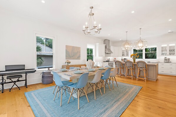 A modern kitchen with a dining area, featuring a piano, a large window, and a chandelier.