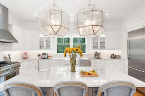 A modern kitchen with white cabinetry, a central island, two hanging light fixtures, and a large window with a view of greenery.