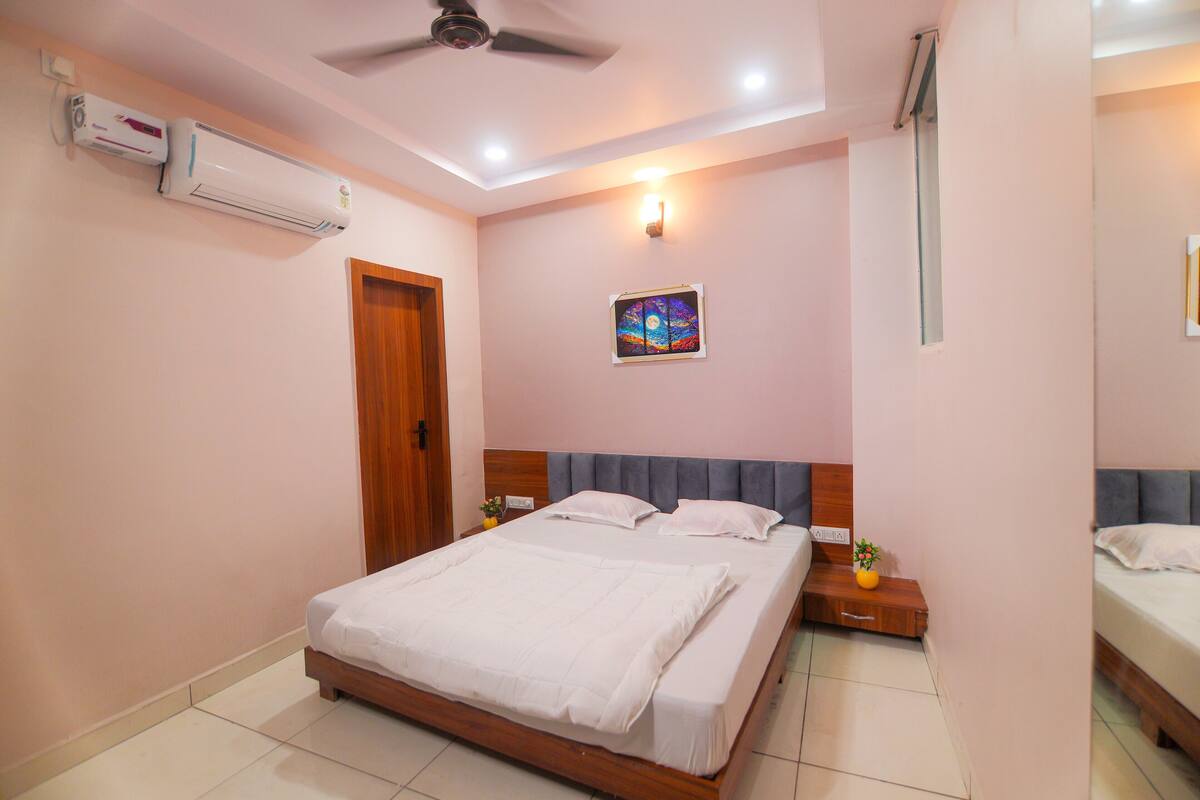 A bedroom with a bed, a ceiling fan, an air conditioning unit, a nightstand, and a wall-mounted picture.