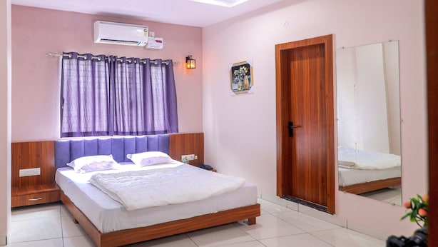 A bedroom with a wooden bed, a nightstand, a wall-mounted air conditioner, a door, and a mirror. - Hotel Dev Krishna Palace (Ujjain)