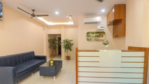 A modern reception area with a sofa, a coffee table, potted plants, and a wooden reception desk. - Hotel Dev Krishna Palace (Ujjain)