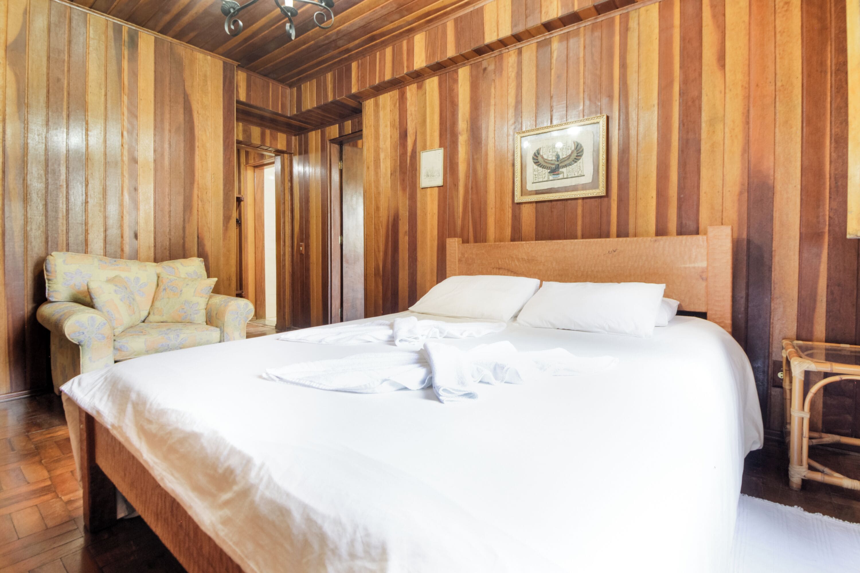 A wooden-paneled room with a large bed, a chair, and a framed picture on the wall.
