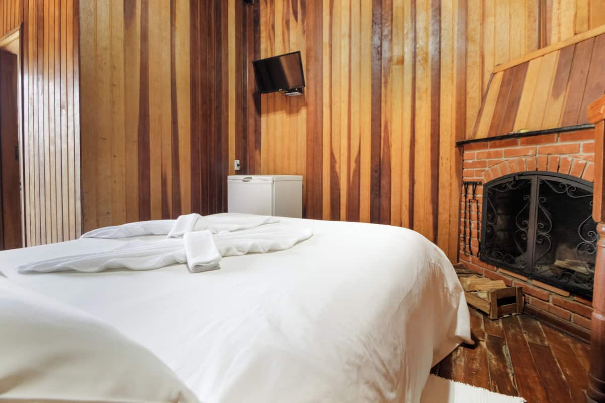 Une chambre d’hôtel avec un mur lambrissé en bois, un lit avec du linge de maison blanc, un téléviseur fixé au mur et une cheminée.