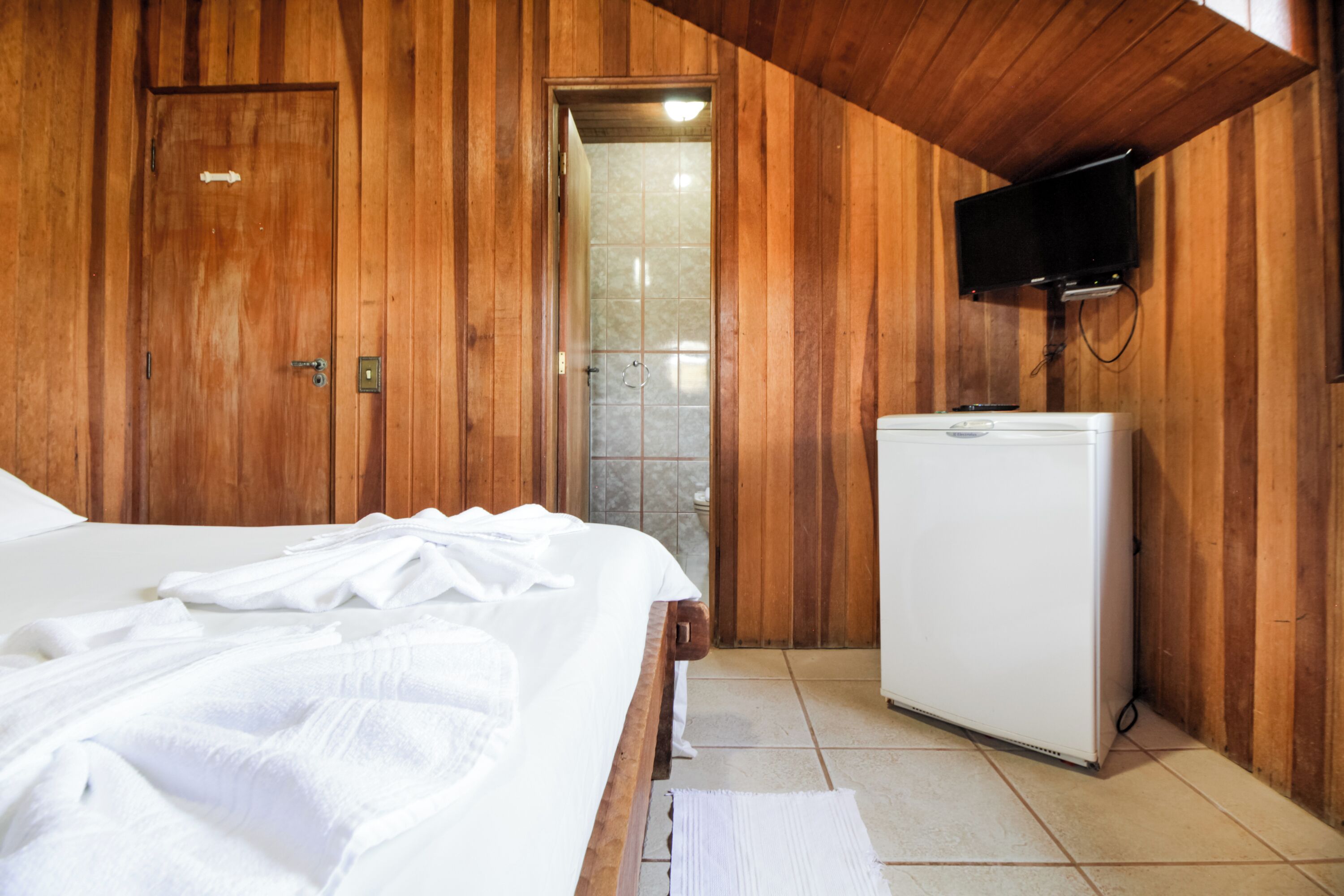 A wooden-paneled room with a bed, a white refrigerator, a television mounted on the wall, and a shower area.