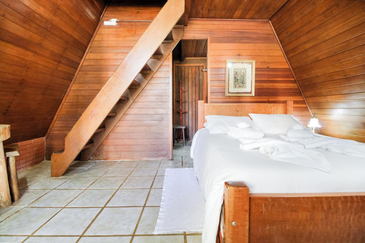 Une chambre avec un plafond en bois à pente, un lit avec du linge de lit blanc, une tête de lit en bois et un tableau encadré au mur.