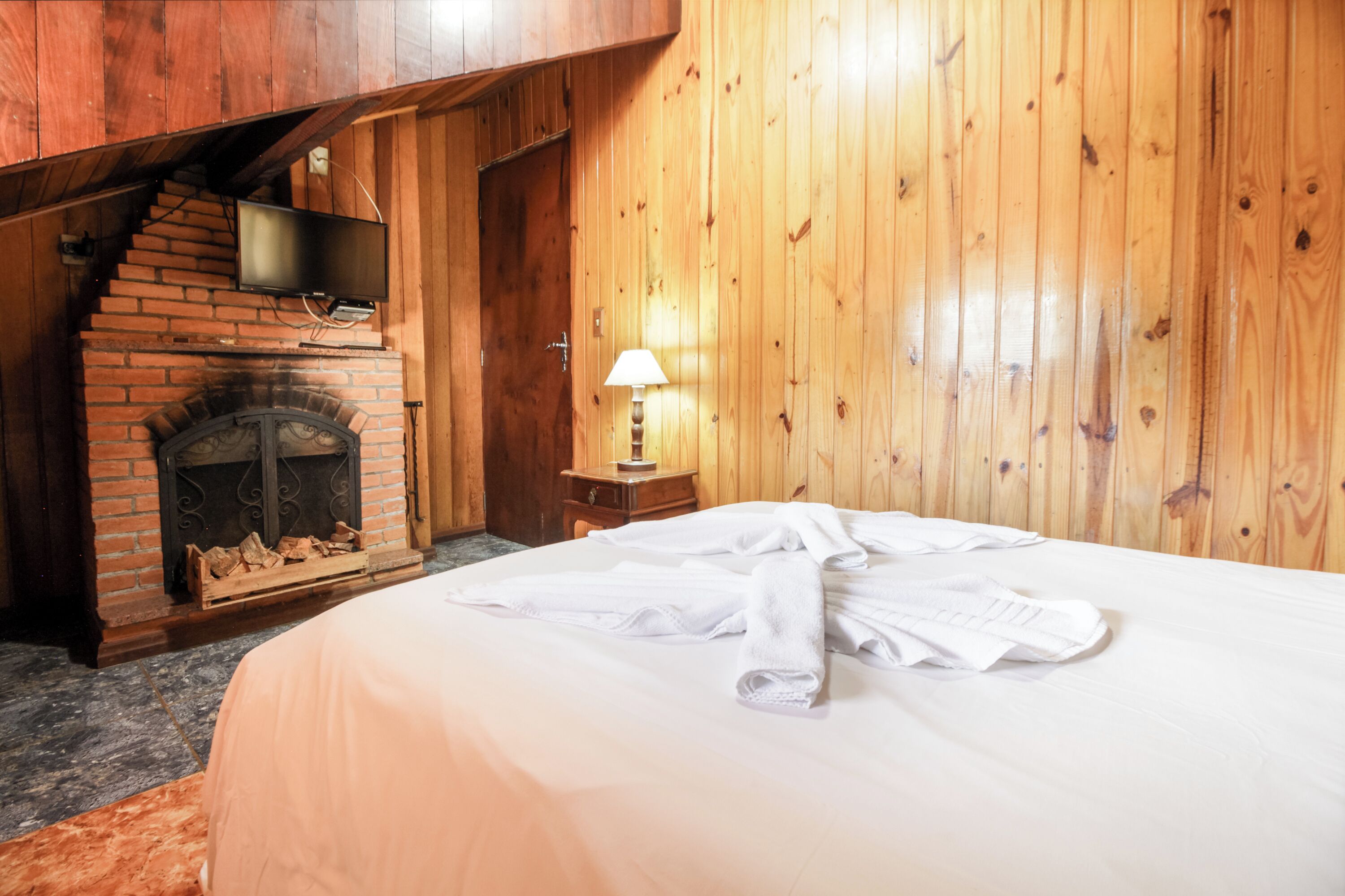 A room with a brick fireplace, wooden walls, a bed with white linens, and a lamp on a nightstand.
