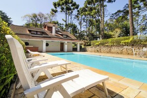 A swimming pool with white lounge chairs, a stone wall, and a house with a tiled roof in the background. - VELINN Pousada Torre Branca (Camanducaia)